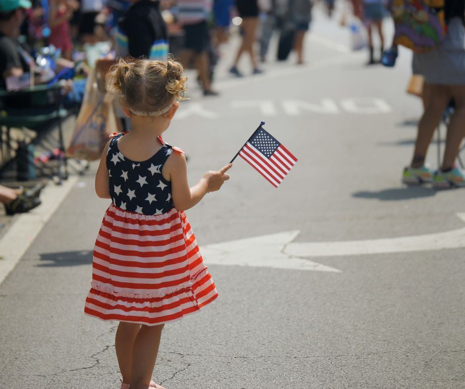 A small child in a red, white, and blue dress, holding an americal flag while watching a parade