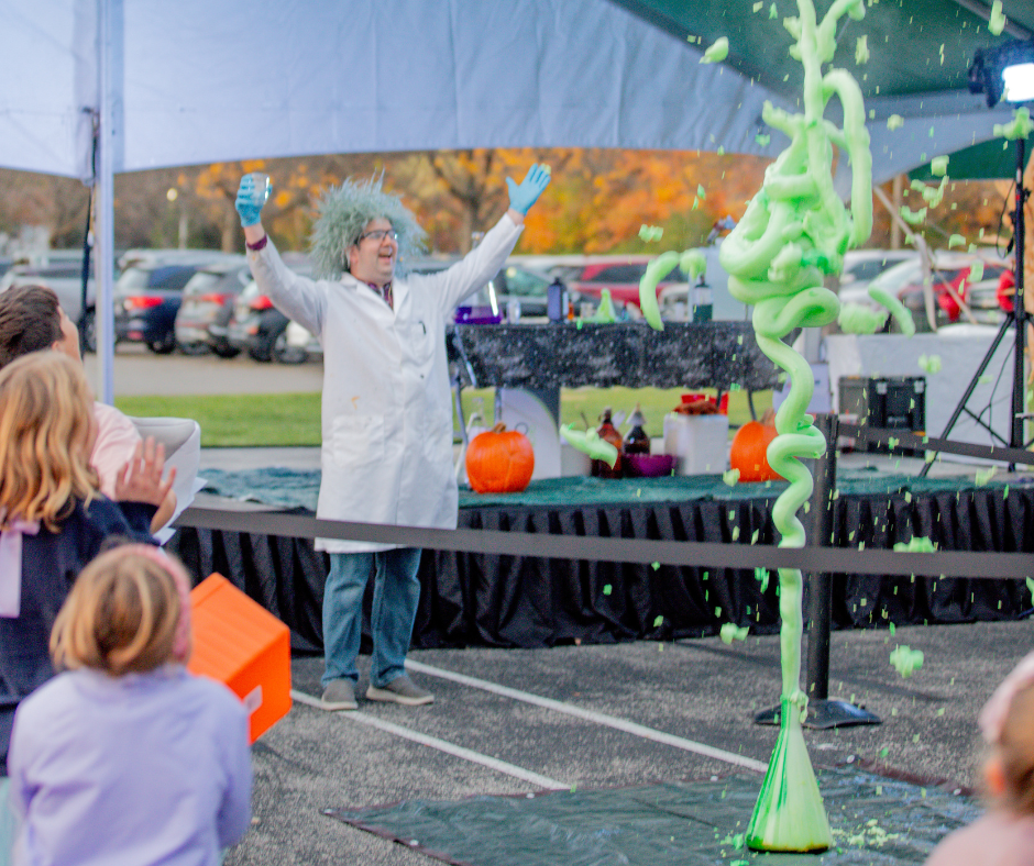 Photo of a man in a scientist costume cheering in front of a crowd as a "chemical bottle" expl