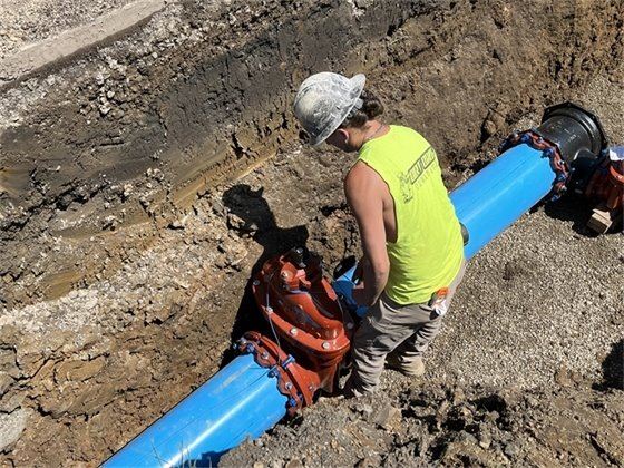 Female construction worker in  dug out pit with large blue pipe, replacing city waterline.
