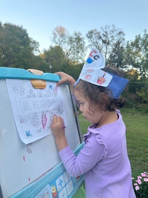 A little girl wearing a paper crown colors a paper on an easel, the paper reads "Hi Neighbor."